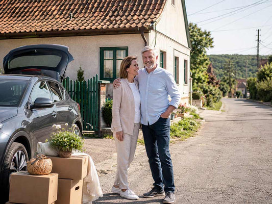 Paar beim Umzug nach Ungarn vor einem Dorfhaus mit Auto und Umzugskartons an einem sonnigen Tag