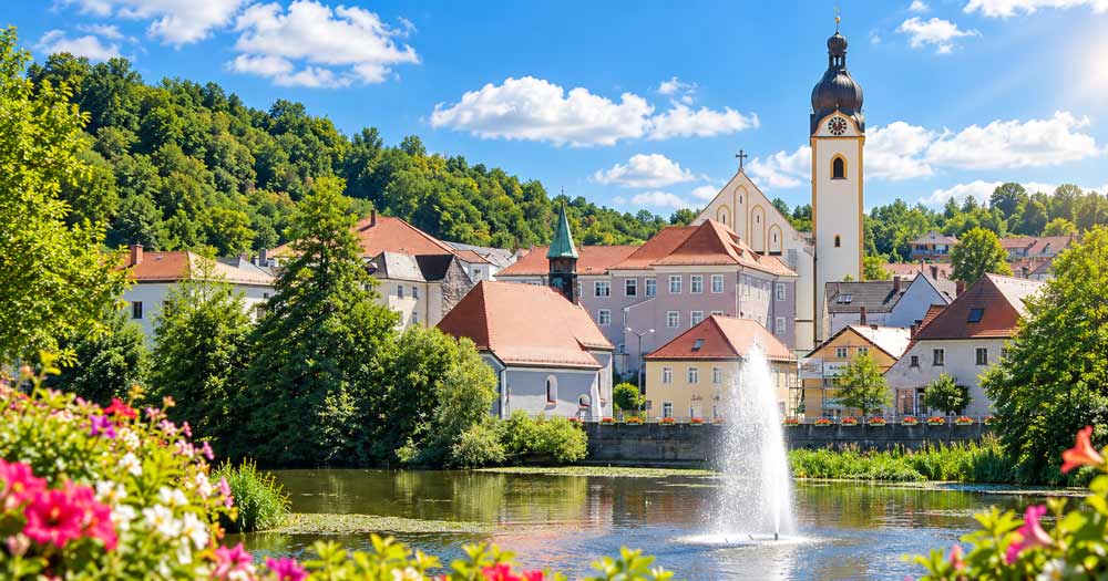 Schwandorf mit Fluss, Springbrunnen, bunten Blumen im Vordergrund, historischer Bebauung und markantem Kirchturm vor grünem Hügel und blauem Sommerhimmel.
